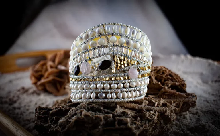 The photo showcases a beautifully crafted bracelet, intricately designed with an array of pearls, beads, and gemstones. The bracelet is prominently displayed on a textured, sandy surface, which appears to be part of a natural or beach-like setting. The background is softly blurred, ensuring the bracelet remains the focal point of the image. The intricate patterns of the bracelet, with its combination of white, gold, and dark-colored beads, create a striking contrast against the earthy tones of the background. The overall composition exudes a sense of elegance and craftsmanship, highlighting the unique beauty of the jewelry piece.