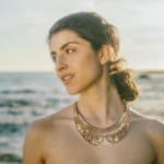 The photo features a young woman standing by the seaside, with the ocean and a blurred shoreline in the background. She is turned slightly to the side, gazing thoughtfully into the distance. The woman has a serene expression on her face, her dark hair is tied back in a loose, low ponytail, and the sunlight casts a warm glow on her skin. She is adorned with a stunning statement necklace made of intricate designs and various beads, adding an elegant touch to her otherwise simple appearance. The overall mood of the photo is tranquil and reflective, capturing a moment of quiet contemplation by the water.